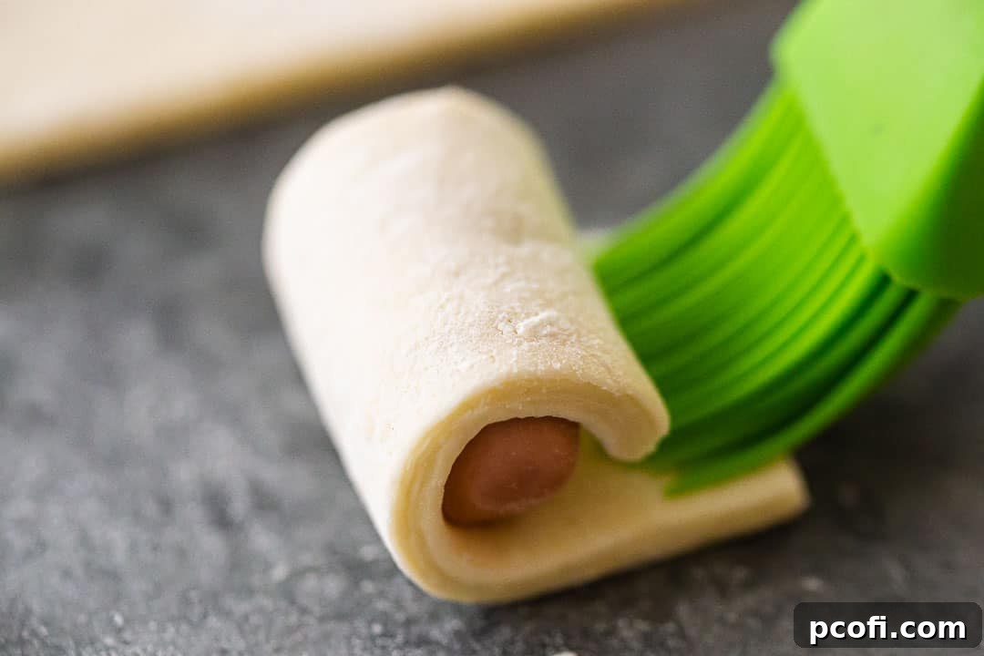 A finger gently pressing the seam of a wrapped hot dog with a drop of water to seal the pastry.