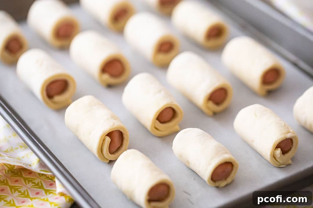 Unbaked pigs in a blanket arranged on a parchment-lined baking sheet, ready for the oven.