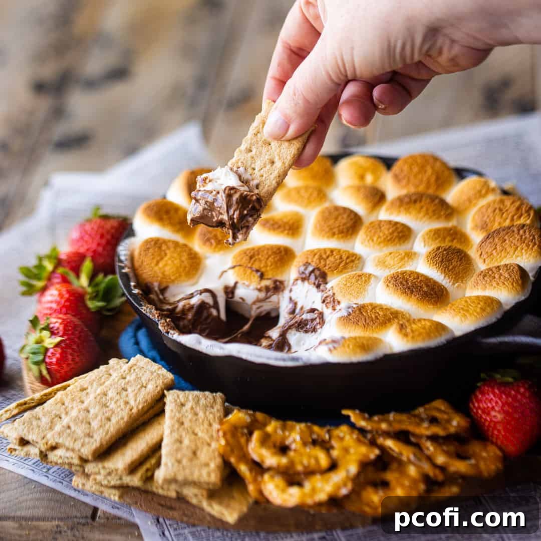 A graham cracker being dipped into a bubbling s'mores dip, freshly baked in a cast iron skillet.