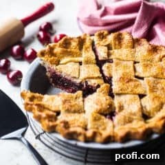 Cherry pie with a few slices cut, on a wire cooling rack with fresh cherries in the background.