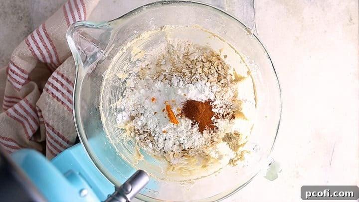 Dry ingredients, including flour, oats, cornstarch, baking soda, salt, cinnamon, and orange zest, being added to the wet mixture in a stand mixer.