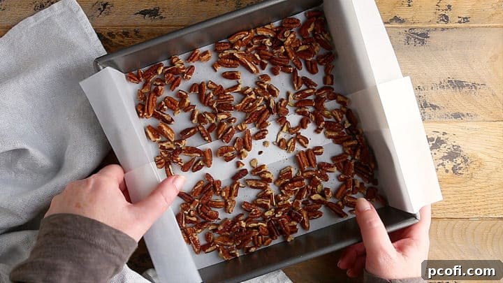 Spreading roughly chopped pecans in an even layer on a parchment-lined baking pan, ready to be toasted in the oven.