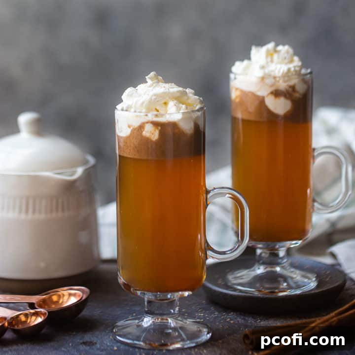 Two tall mugs of hot buttered rum, garnished with whipped cream and nutmeg, with a steaming teakettle in the background, signaling warmth and comfort.