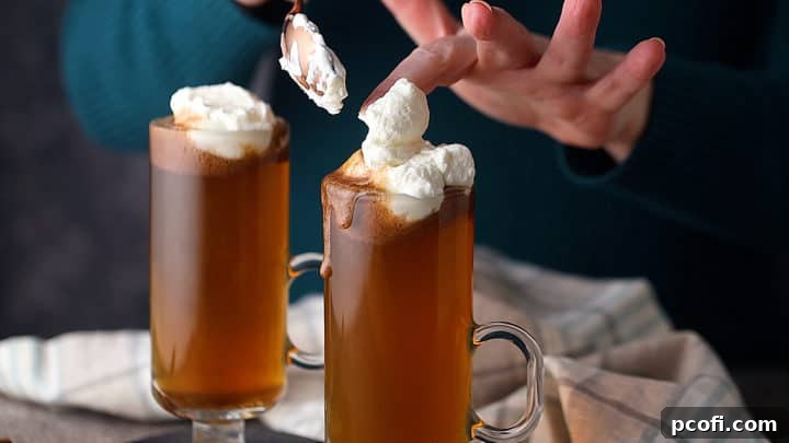 Two mugs of hot buttered rum being topped with fluffy sweetened whipped cream, ready for a final garnish of nutmeg.