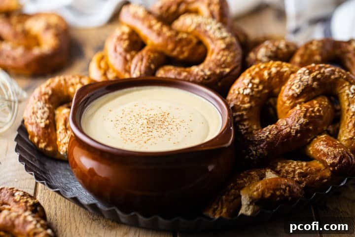 A pot-bellied brown bowl filled with the golden beer cheese dip, perfectly prepared and ready to serve, with a blurred background of soft pretzels.