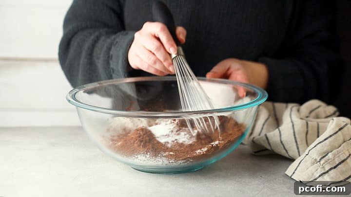 Whisking dry ingredients together in a large glass bowl, including flour, sugar, cocoa powder, baking soda, and salt.