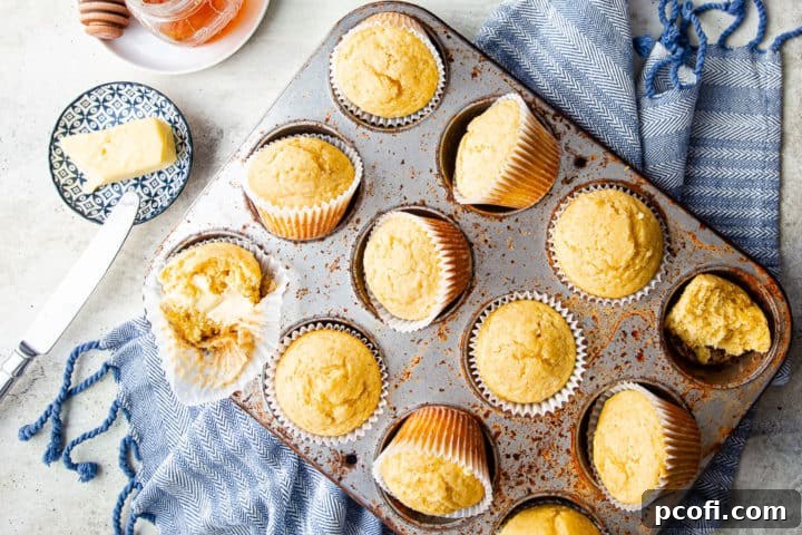 A selection of warm cornbread muffins arranged on a blue kitchen towel, accompanied by a small bowl of soft butter and a jar of honey, inviting a delicious meal.