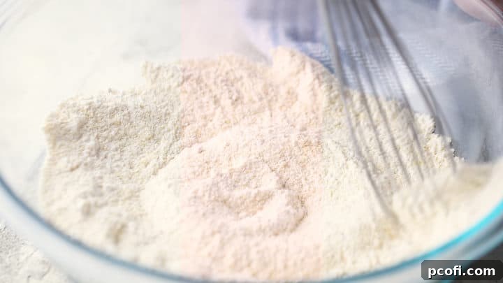 A hand whisking together the dry ingredients (flour, sugar, cornmeal, baking powder, salt) in a large glass bowl.