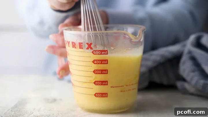 A whisk blending milk, oil, and eggs with slightly cooled melted butter in a large liquid measuring cup.
