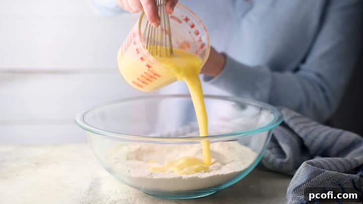 Liquid ingredients being poured from a measuring cup into a large bowl containing dry ingredients for cornbread muffins.