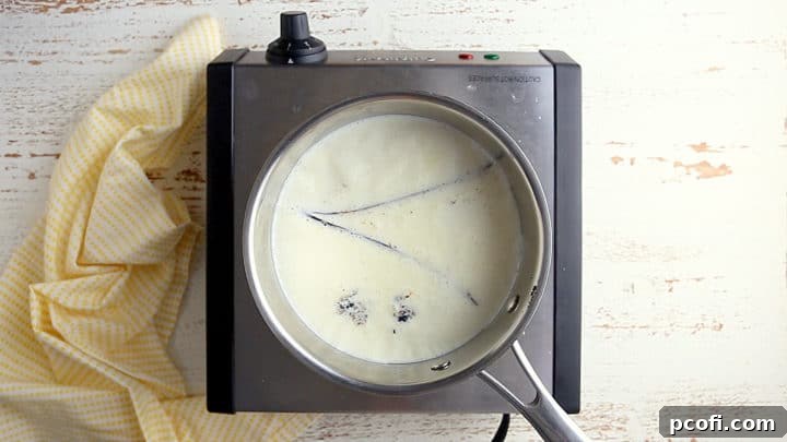 Milk, heavy cream, and a vanilla bean with its scraped seeds are gently heated in a small pot until steam rises, indicating a perfect infusion.