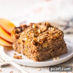 A square of baked oatmeal, served on a white plate with apple slices and honey.