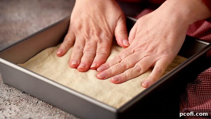 Hands pressing Detroit-style pizza dough into a prepared square pan, ensuring even coverage to the edges.