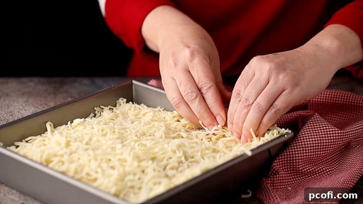 Close-up of hands pressing shredded cheese firmly up the sides of a partially baked Detroit-style pizza crust in a pan, creating the frico edge.