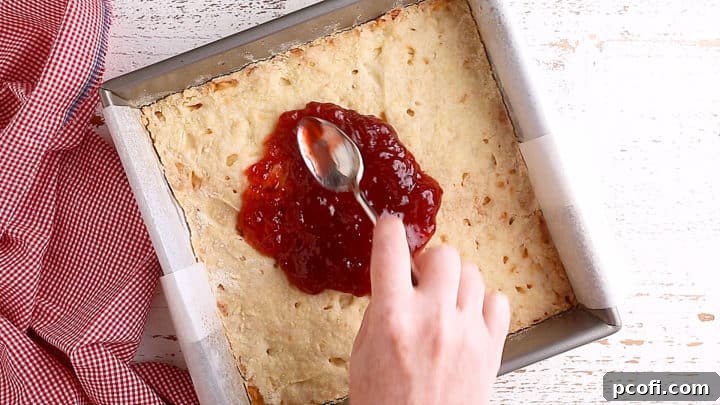Carefully spreading a layer of sweet strawberry jam over the partially baked shortbread crust.