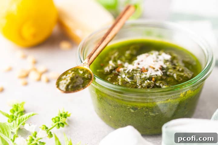A jar of basil pesto on a pale background, surrounded by fresh basil, lemon, parmesan, and pine nuts.