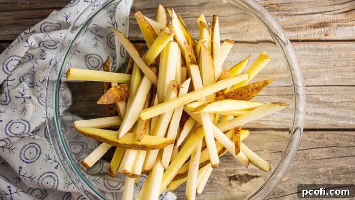 Freshly cut, uncooked potato planks and batons in a large clear glass bowl, ready for soaking.