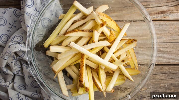 Uncooked potato fries tossed with oil, cornstarch, and salt in a large glass bowl, ready for baking.