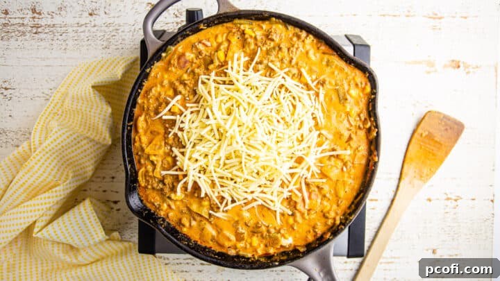 Shredded sharp cheddar cheese being added to the hot and creamy Mexican corn dip mixture in a skillet, ready to be stirred until melted and gooey.