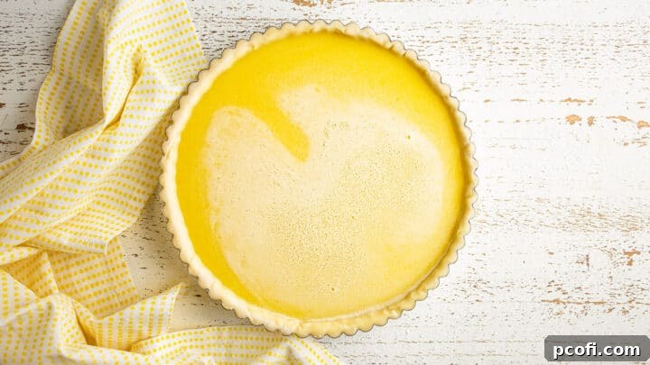 Overhead view of an unbaked lemon tart, with its vibrant yellow filling already poured into the crust-lined pan, resting on a charming yellow checked cloth.