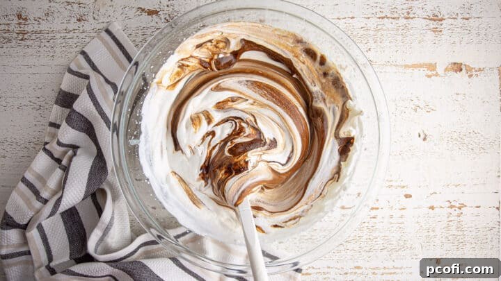 A silicone spatula gently folding whipped cream, condensed milk, and espresso mixture together in a large bowl, creating a harmonious no-churn ice cream base.