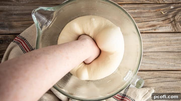 A hand gently punching down a large, risen ball of pretzel bun dough in a bowl.