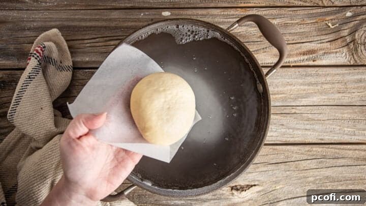 A shaped pretzel bun gently placed into a pot of simmering baking soda water.