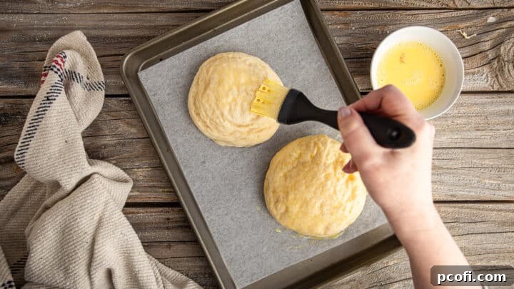 Boiled pretzel buns being brushed with egg wash on a parchment-lined baking sheet.