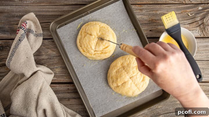 A baker scoring the top of an unbaked pretzel bun with an 'x' shape using a baker's lame.