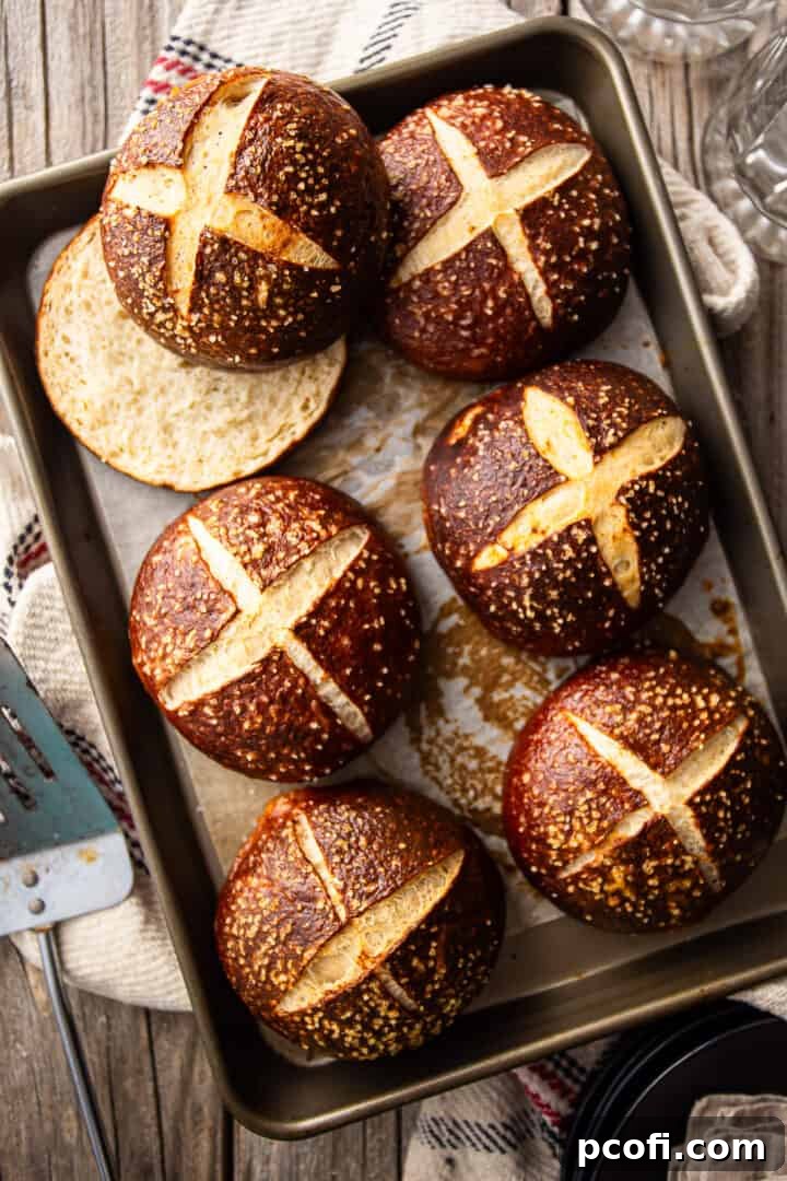 Overhead shot of freshly baked pretzel buns presented on a parchment-lined baking sheet, accompanied by beer glasses and a grill spatula.