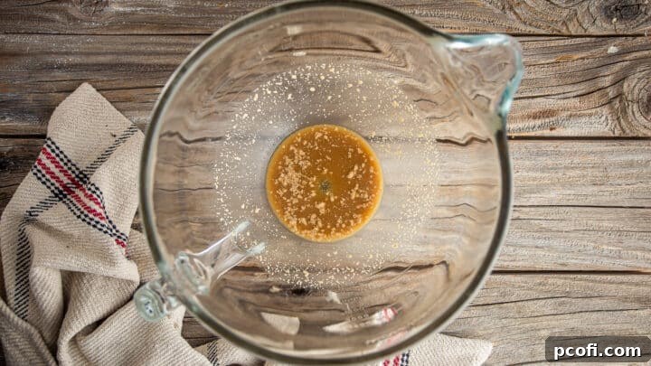 Active dry yeast being added to the sugar water mixture for pretzel buns, showing the initial stage of activation.