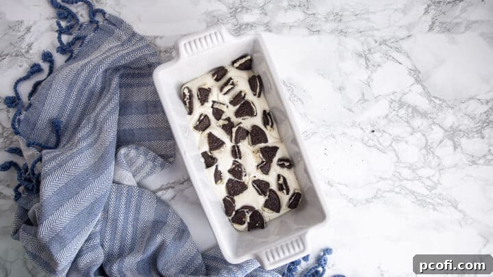 Layering crushed Oreo cookies and no-churn ice cream mixture into a white ceramic loaf pan.