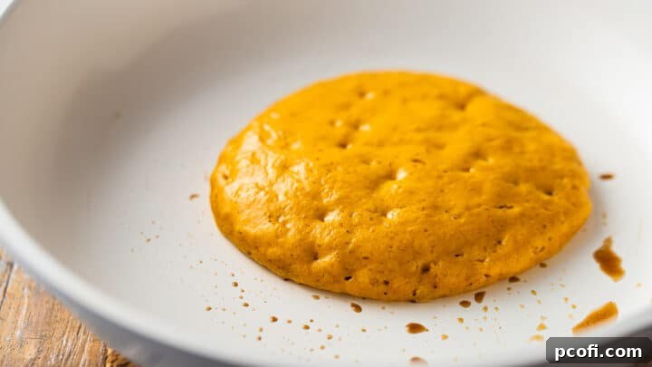 Pumpkin pancake cooking in a skillet, ready to flip.