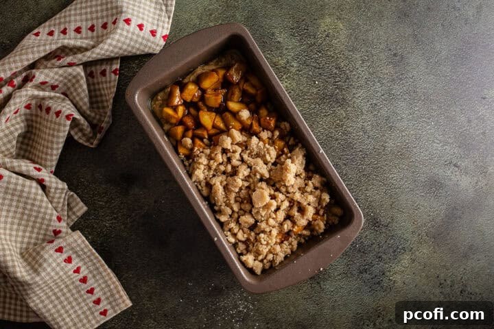 Layers of cooked apples and cinnamon crumb topping meticulously placed over the initial layer of bread batter in a loaf pan.