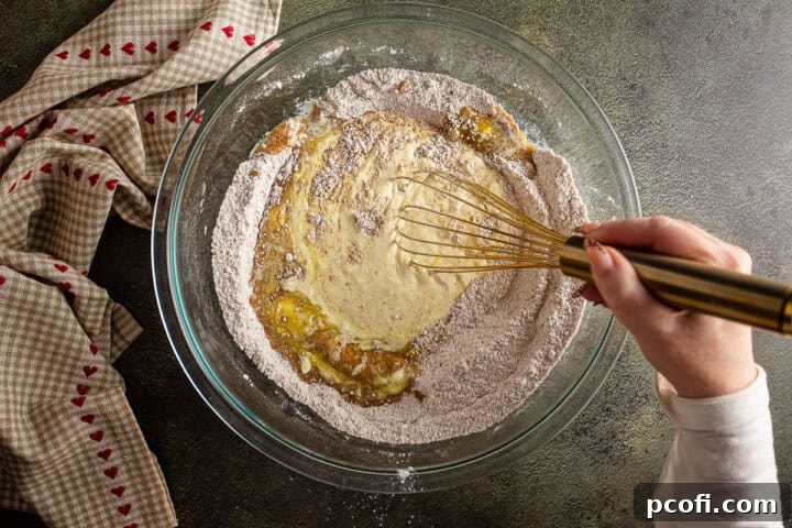 The process of gradually incorporating dry ingredients into the wet mixture in the bowl, ensuring a well-blended apple bread batter.