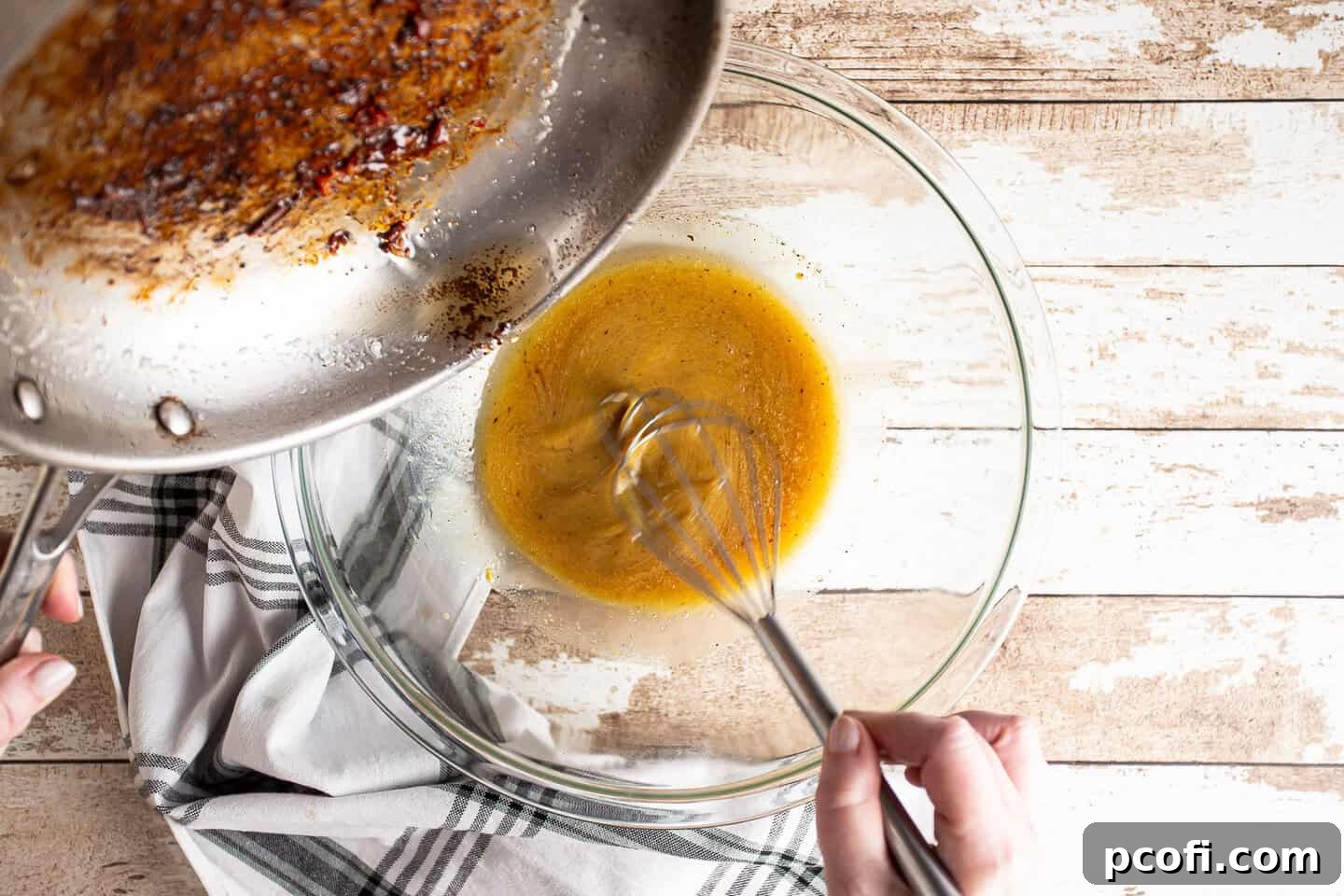 A hand whisking hot bacon fat into a bowl containing vinaigrette ingredients, creating a smooth emulsion.