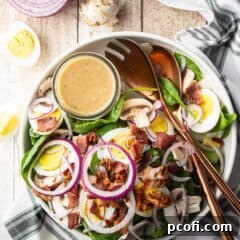 Spinach salad with warm bacon dressing, served in a white ceramic bowl with copper salad serving utensils, highlighting its fresh ingredients.