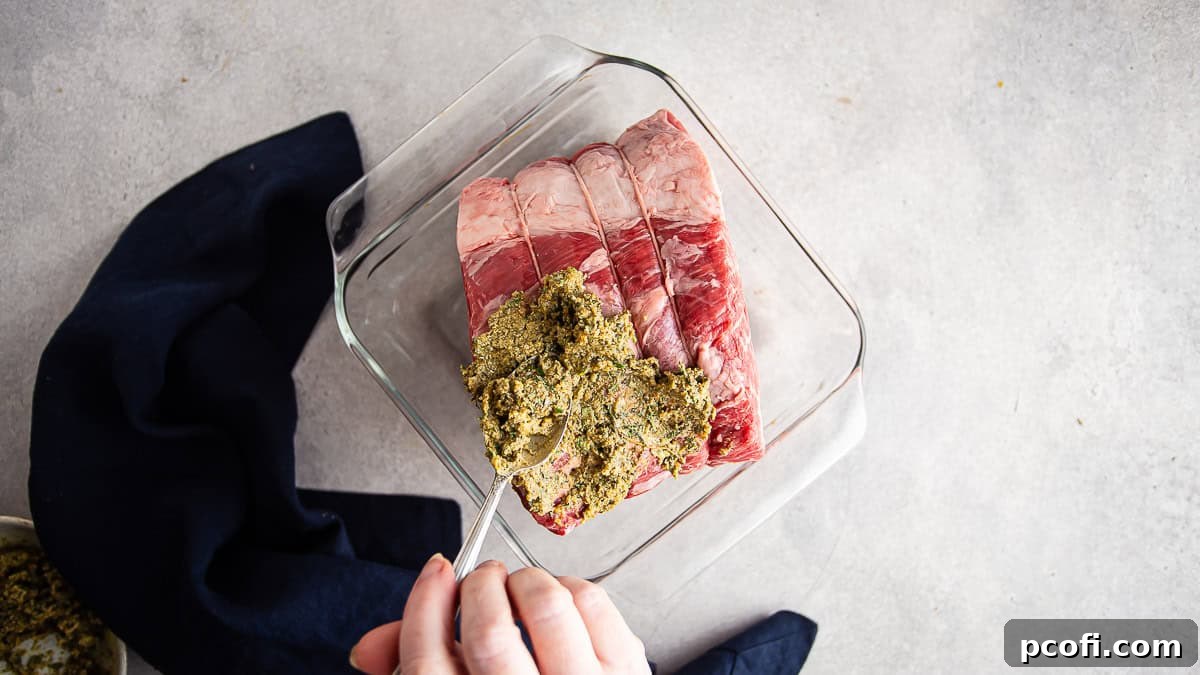 A skilled hand is spreading the fragrant herb paste evenly over the majestic standing rib roast, ensuring every part is covered in a glass roasting pan.