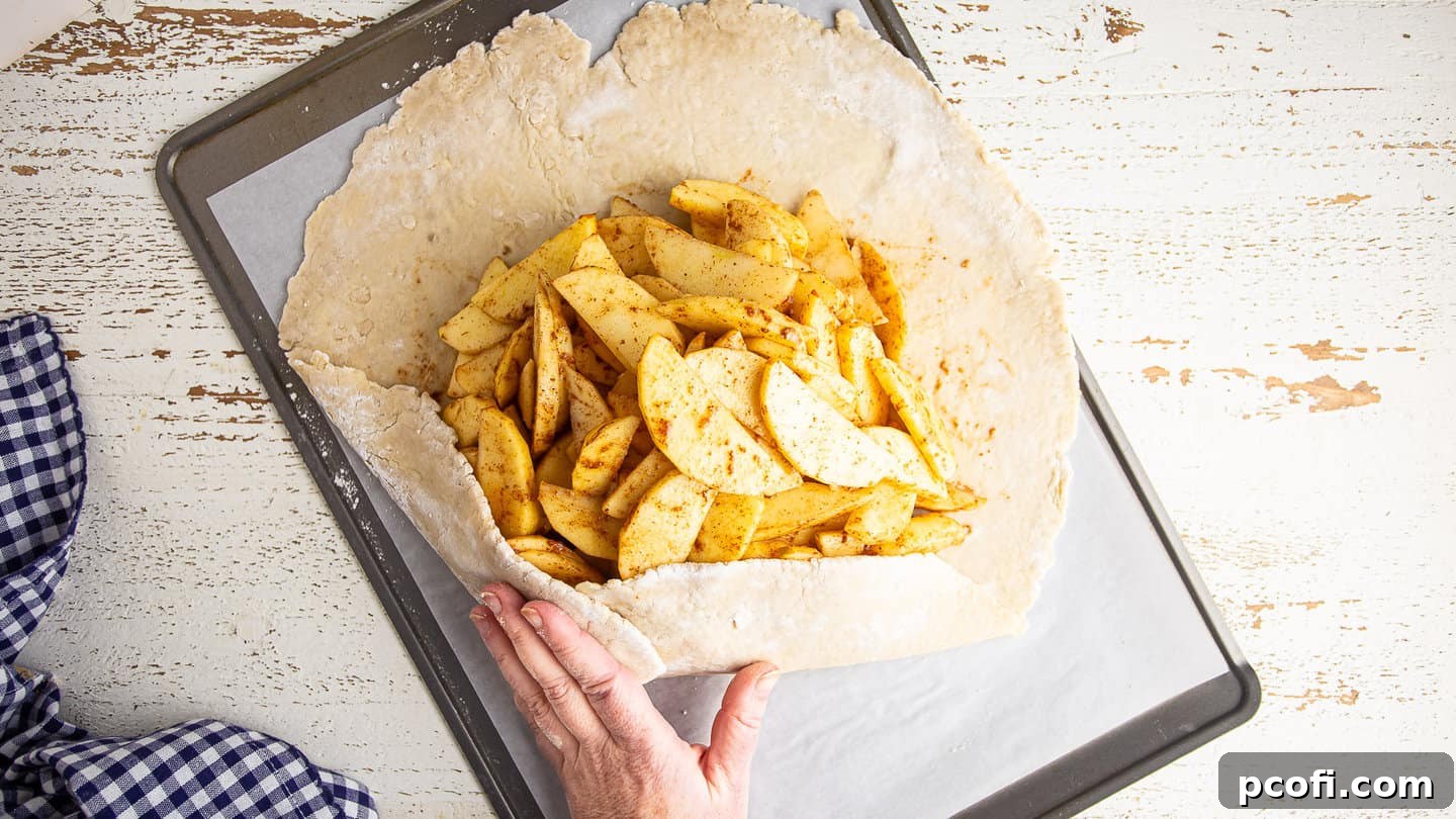 Hands carefully forming an apple galette on a baking sheet, folding the pastry edges over the filling.