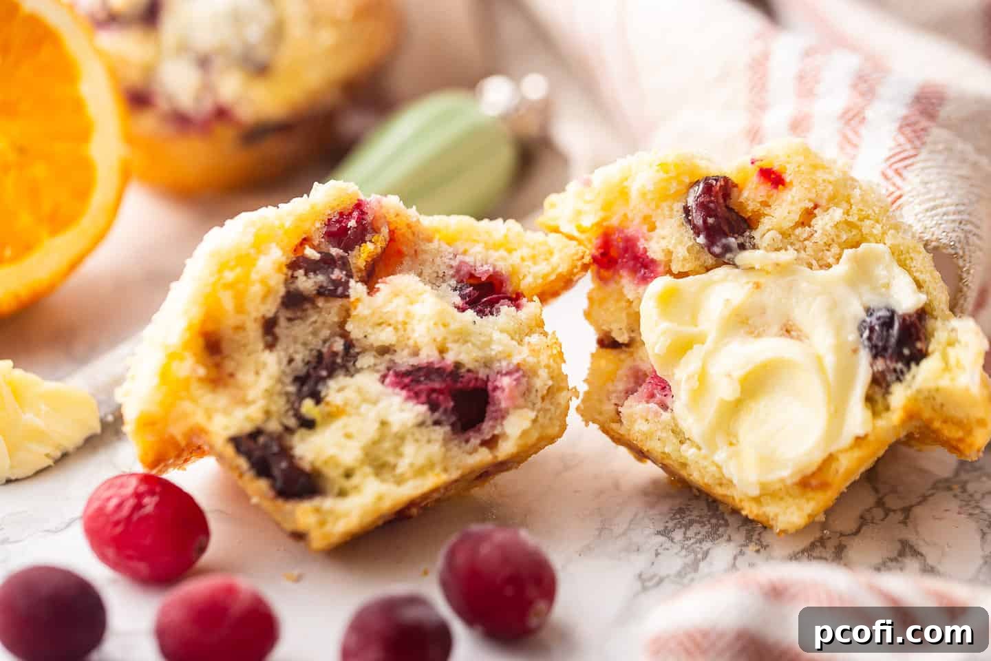 A delectable cranberry orange muffin, gently buttered, with a fresh orange and a red striped kitchen towel completing the cozy, inviting scene.