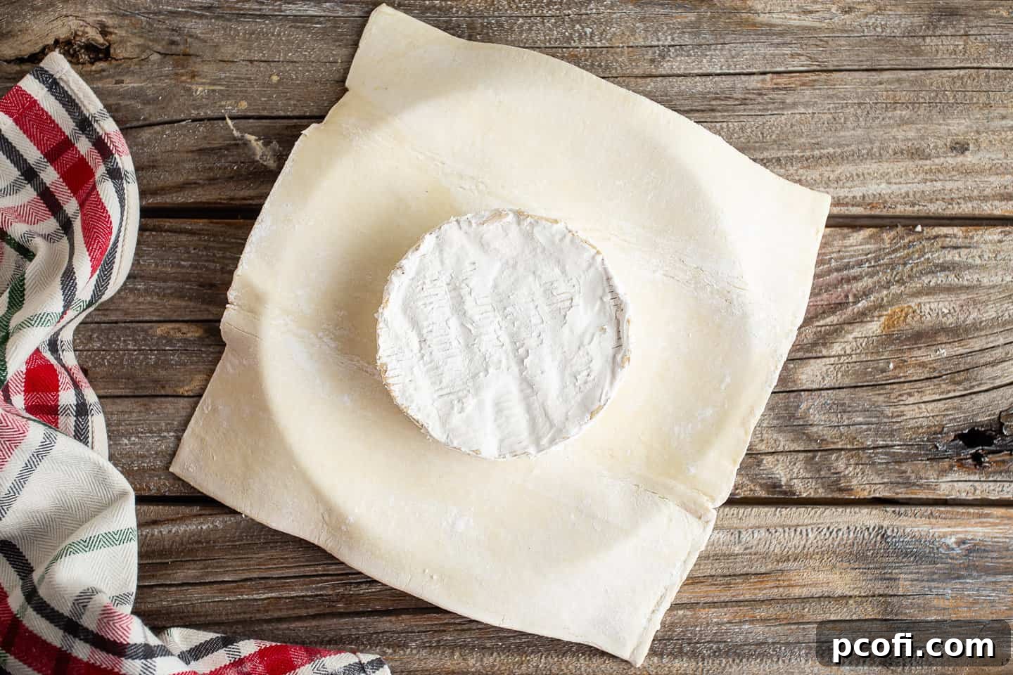 A perfectly round wheel of brie cheese positioned in the center of a square sheet of puff pastry, ready for topping.