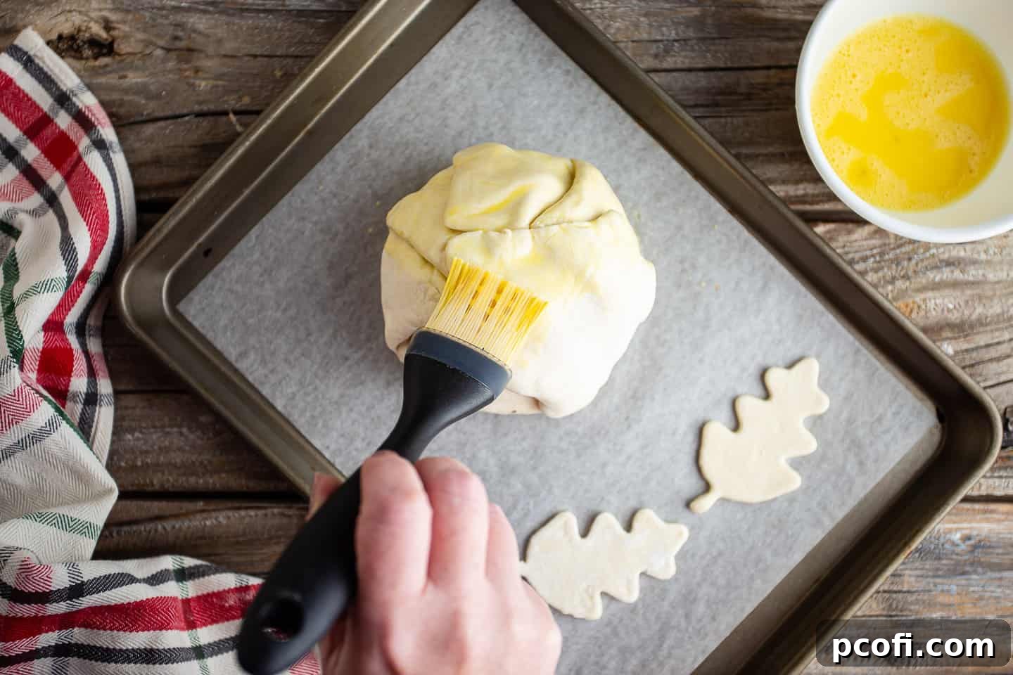 A pastry brush applying egg wash over the surface of the assembled brie en croute, preparing it for baking.