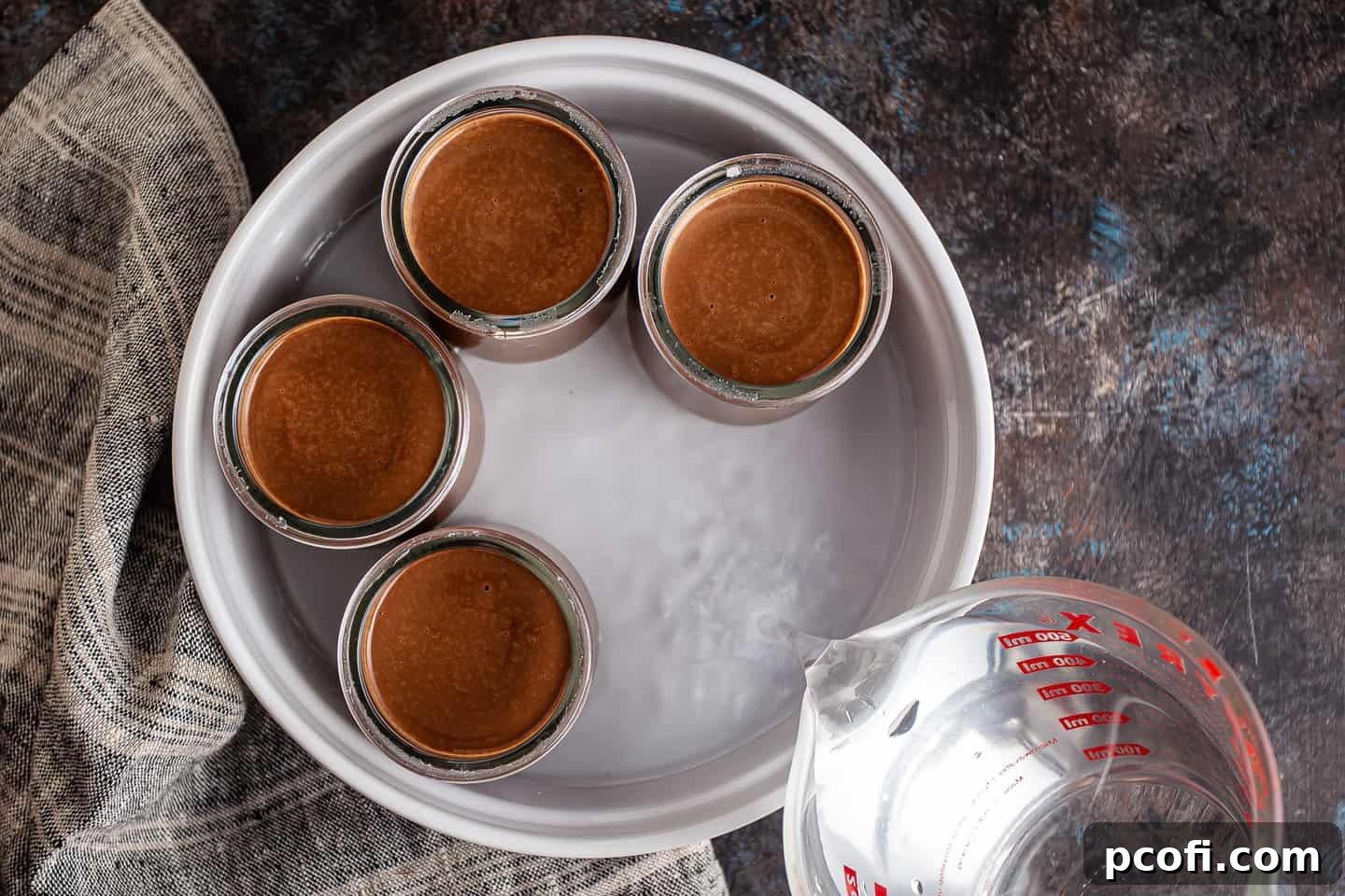 Hot water being carefully poured into a larger baking dish, surrounding individual ramekins filled with pot de creme custard, establishing the essential water bath.