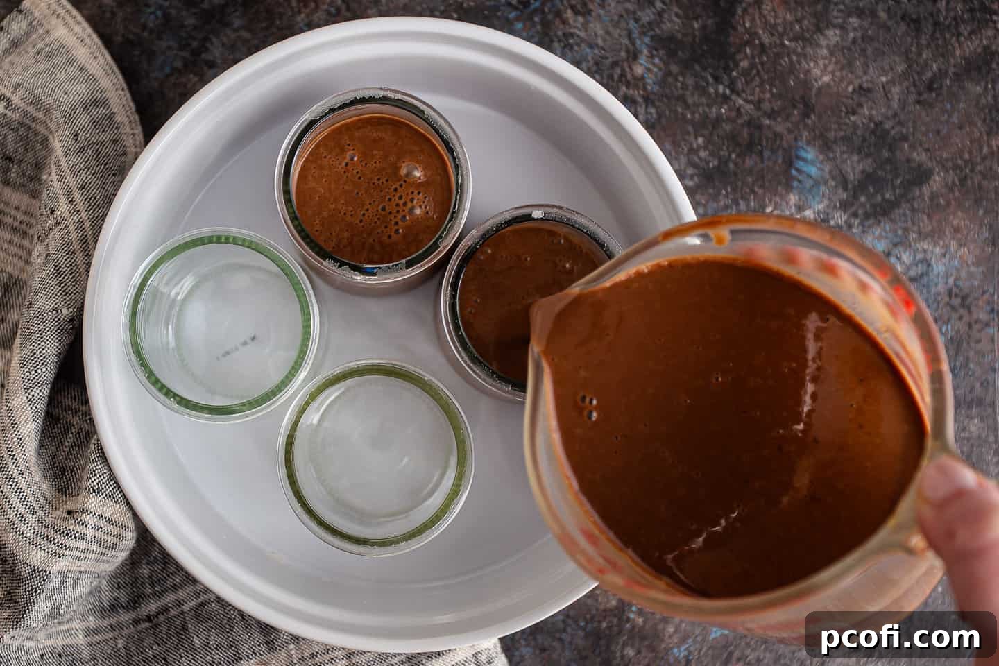 The chocolate custard mixture being carefully poured into individual serving dishes, which are placed within a larger baking pan, in preparation for the water bath.