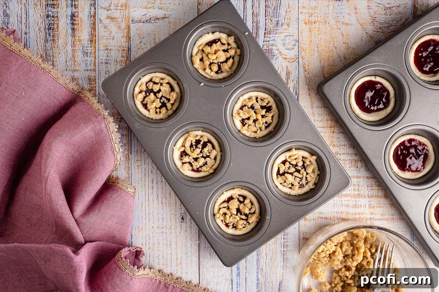 Unbaked raspberry tarts with jam filling, generously topped with streusel, arranged in a cupcake pan.