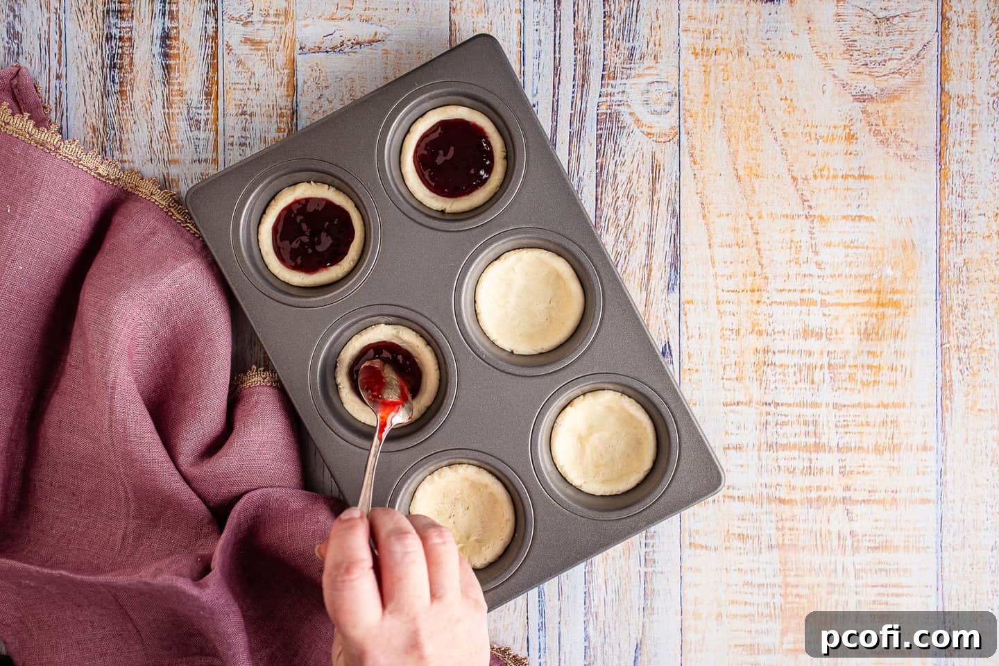 Filling individual pastry shells with bright red raspberry jam using a spoon.