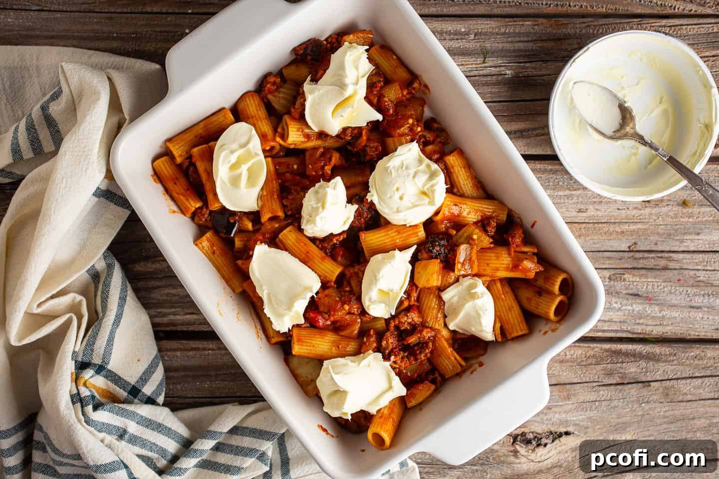The process of dropping dollops of creamy mascarpone cheese over a layer of rigatoni pasta in a baking dish.