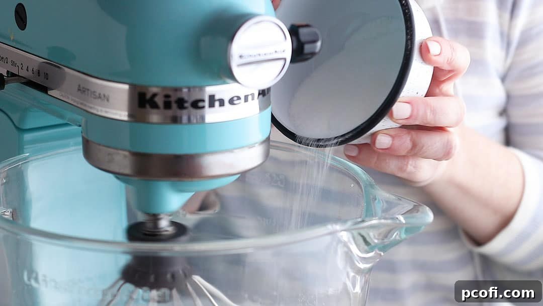 Granulated sugar and cornstarch being slowly streamed into whipping meringue, ensuring proper dissolution for a stable, no-weep topping.