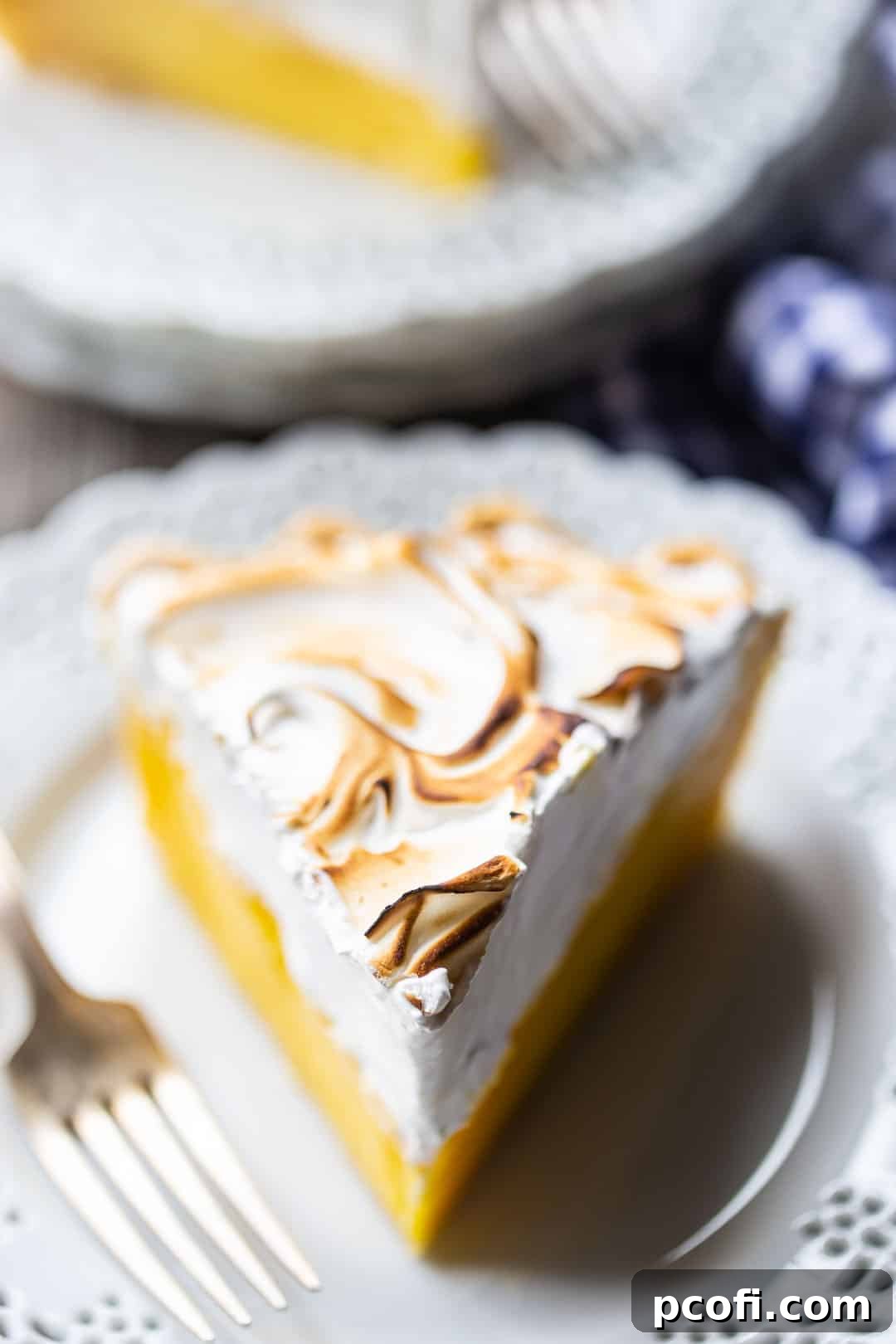A perfectly sliced wedge of lemon meringue pie presented on a white plate, with a blue checkered cloth in the background, highlighting its layers.