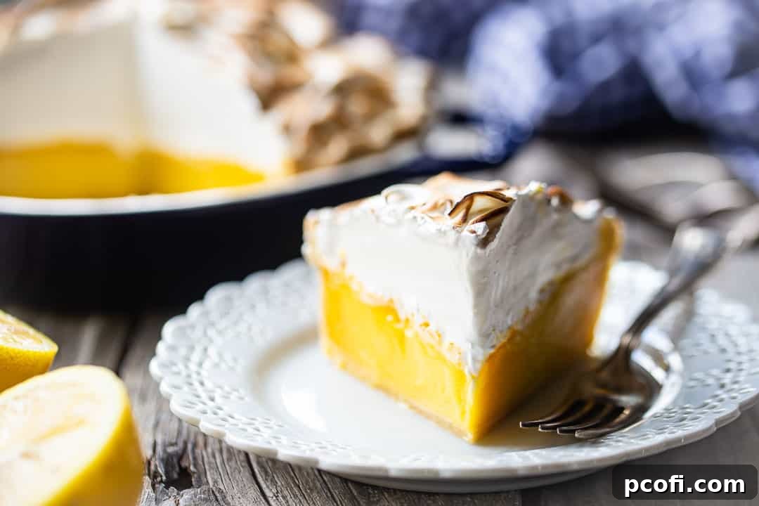 A generous slice of easy lemon meringue pie on a plate, with a soft blue checked cloth and a whole lemon in the background.